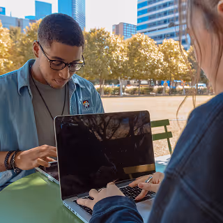 Young man and woman seated at a green table in an outdoor urban park, both focused on their laptops.