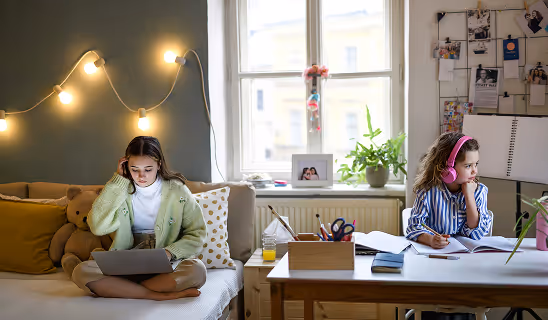 One girl sits on a bed using a laptop while the other sits at a desk wearing headphones and writing, with warm string lights in the background.