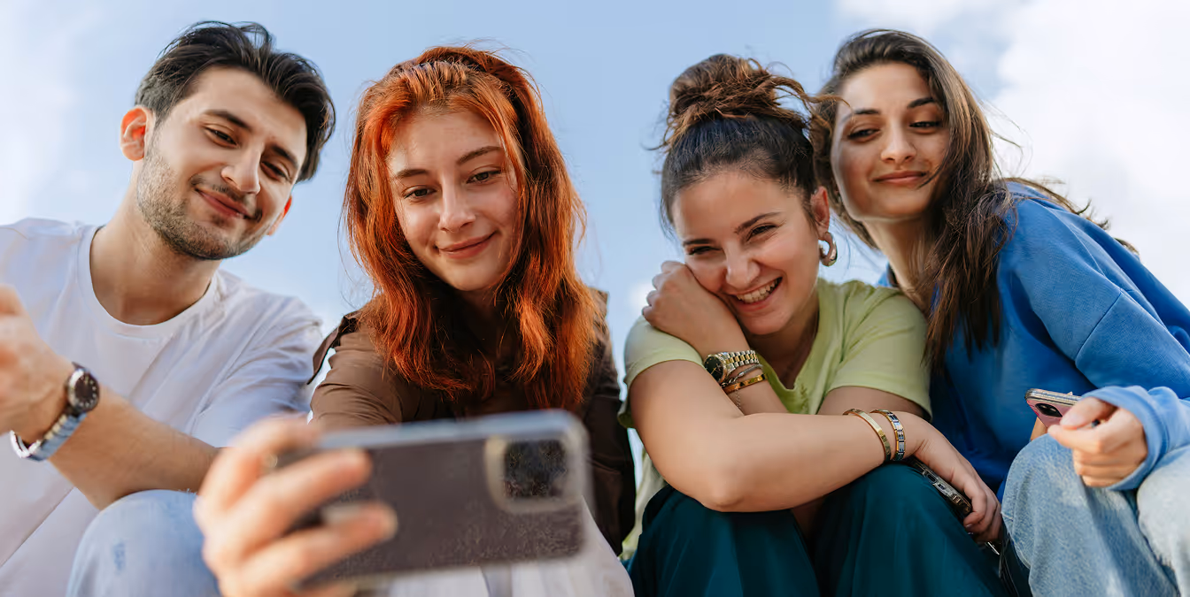 Young adults sitting close together outdoors, smiling at a smartphone camera.