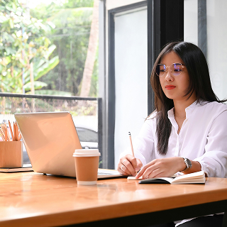Professional woman in glasses writing in a notebook while working on a laptop in a bright, modern office with a coffee cup nearby.