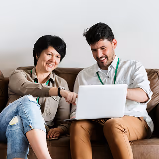 Smiling man and woman sitting on a couch, reviewing something on a laptop screen.