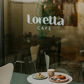 Café window with the words “Loretta Café” and a feather graphic, with coffee and pastries on a table outside.