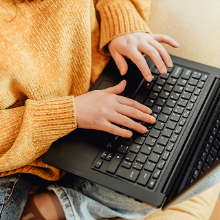 Close-up of a person in a mustard sweater and jeans typing on a black laptop while sitting comfortably indoors.