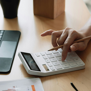 A close-up of a person using a white calculator with a pencil in hand, with a laptop and papers on a wooden desk.
