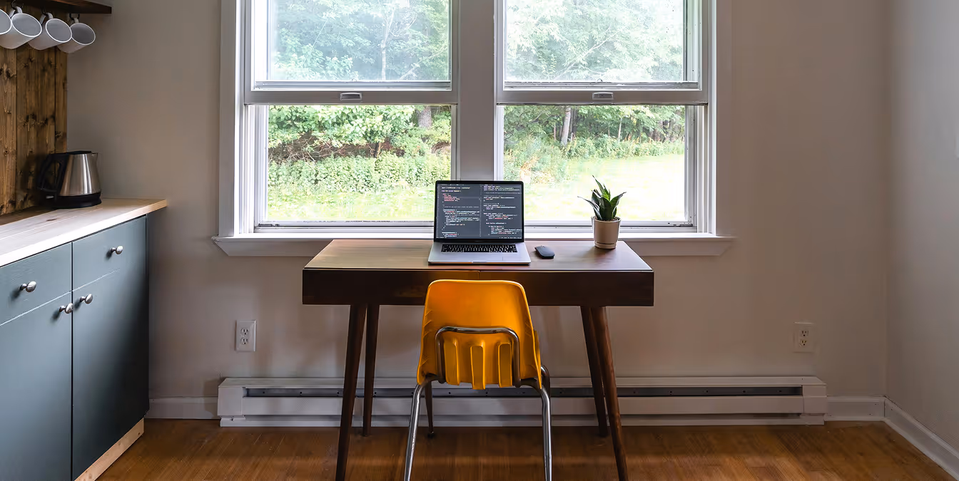 A minimalist home office with a wooden desk, laptop displaying code, a yellow chair, a small plant, and a window overlooking greenery.