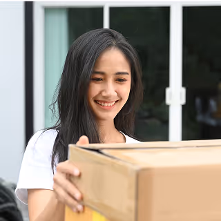 A smiling woman holds a large cardboard box outside her home, appearing pleased with the delivery.
