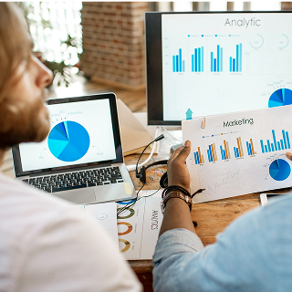 Two people review printed marketing analytics charts while also viewing bar and pie graphs on laptops and monitors.