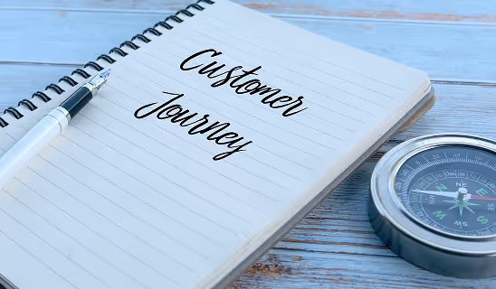 A notebook with “Customer Journey” written in cursive, next to a pen and a compass on a wooden surface.