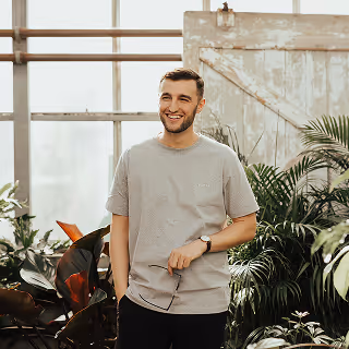 A man in a gray t-shirt stands in a greenhouse surrounded by lush plants, smiling and holding his glasses.