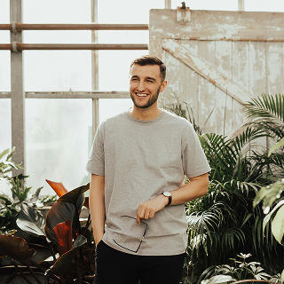 A man in a gray t-shirt stands in a greenhouse surrounded by lush plants, smiling and holding his glasses.