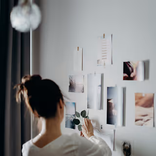 A woman pins photographs and notes to a wall, creating a mood board or inspiration collage.