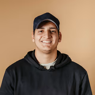 A smiling man wearing a black hoodie and a navy baseball cap, standing against a beige background.