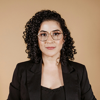 A woman with short curly hair wearing glasses and a black blazer, standing against a beige background and looking confidently at the camera.