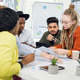 A diverse group of four people gathered around a table in an office, reviewing printed charts and documents during a discussion.