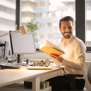 Smiling man in a white shirt sitting at a bright office desk, holding an open book while working at a computer.