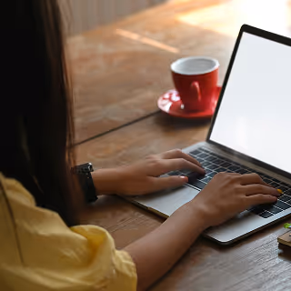 Person in a yellow shirt typing on a laptop at a wooden table with a red coffee cup nearby.