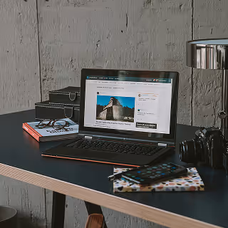 Laptop on a modern desk displaying a travel or lifestyle blog, with a camera, books, and a lamp in the background.