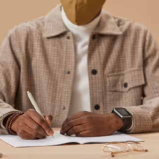Close-up of a man wearing a beige checkered jacket and face mask, writing in a notebook at a desk with glasses placed beside him.