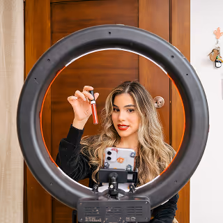 A woman holding a lipstick tube, speaking into a phone camera set up with a ring light for recording beauty content.
