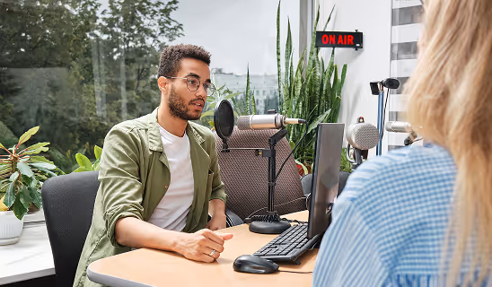 A man wearing glasses speaks into a microphone in a studio while another person listens, with an “On Air” sign in the background.