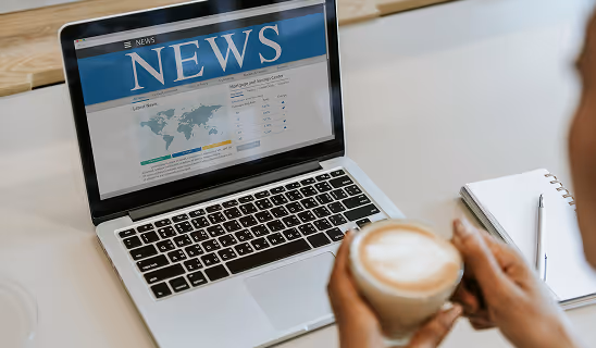 Close-up of a laptop screen displaying a news website while a person holds a coffee cup and sits at a desk with a notebook and pen.
