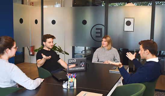 Four colleagues having a discussion around a conference table in a modern office space with glass walls.