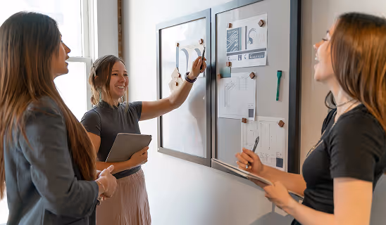 Three women smiling and collaborating while pointing to design boards during a team meeting.