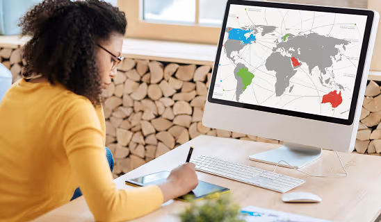 Woman working at a desk analyzing a world map with highlighted countries on a desktop monitor.