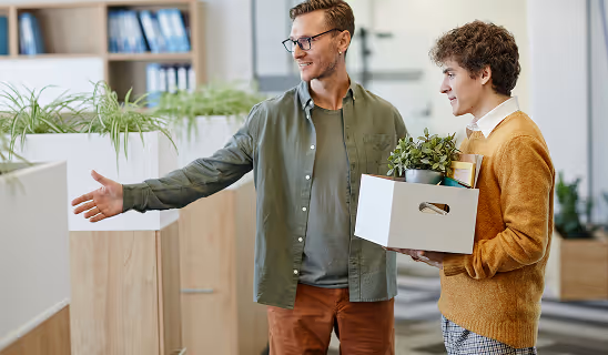Smiling man greeting a newcomer holding a box of personal items in an office environment.