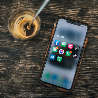Smartphone on a wooden table next to a glass of coffee, showing a “Social” folder with popular social media app icons like Facebook, Instagram, TikTok, and WhatsApp.