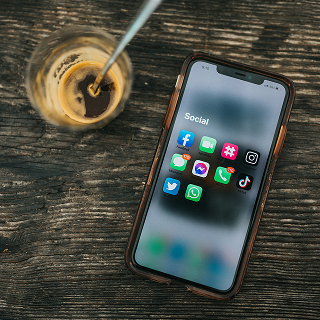 Smartphone on a wooden table next to a glass of coffee, showing a “Social” folder with popular social media app icons like Facebook, Instagram, TikTok, and WhatsApp.