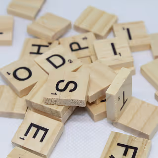 Pile of wooden Scrabble tiles scattered on a white surface, displaying various letters.