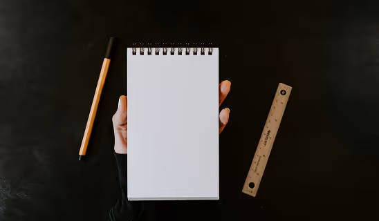 Hand holding an empty spiral-bound notepad, with a pen and ruler placed on a dark tabletop.