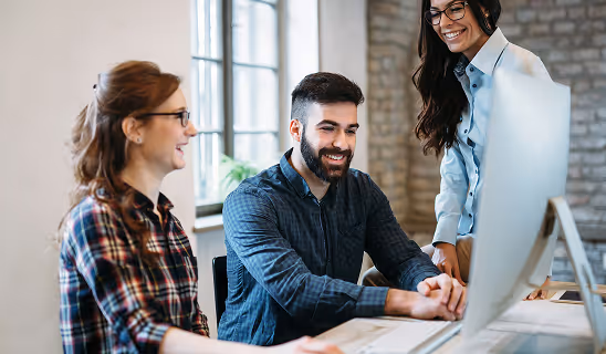 Group of three people collaborating at a computer in a modern office space, smiling and discussing ideas.