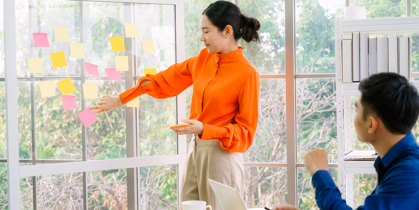 Woman in an orange blouse pointing to sticky notes on a glass wall during a brainstorming session with a seated male colleague.