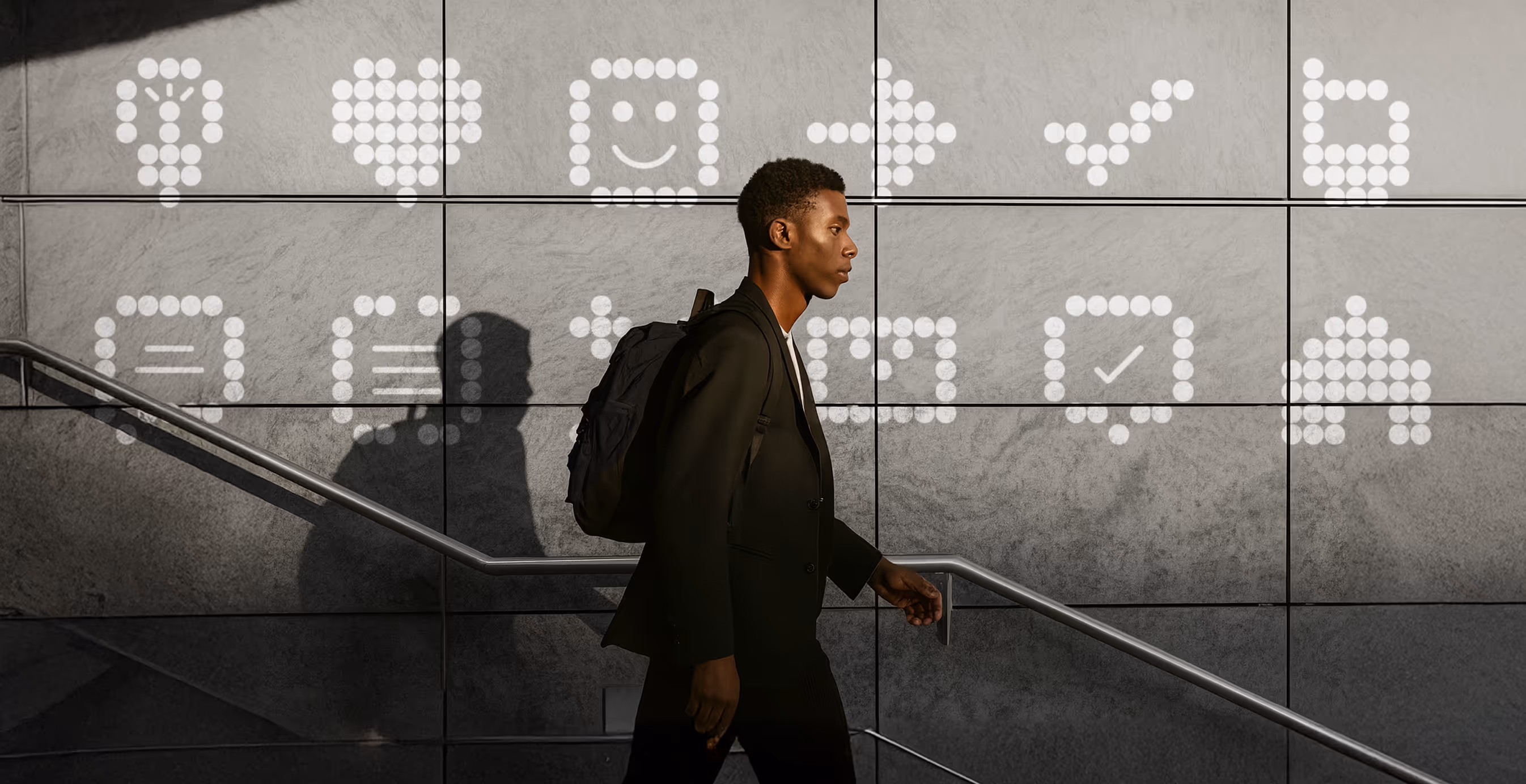A man with a backpack walks past a wall of large illuminated dot-matrix icons, including a light bulb, heart, and check mark, projected in white light against gray tiles.