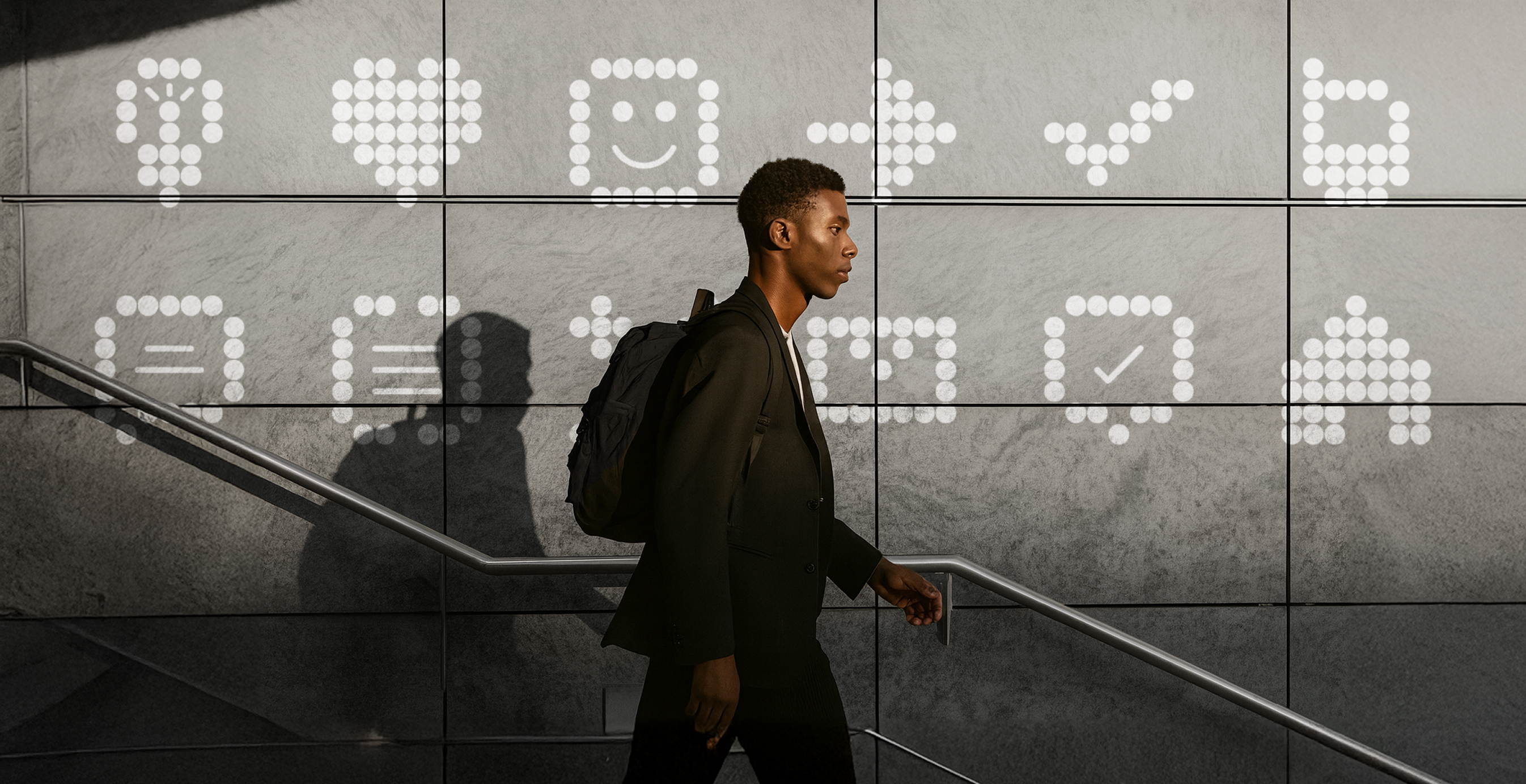 A man with a backpack walks past a wall of large illuminated dot-matrix icons, including a light bulb, heart, and check mark, projected in white light against gray tiles.