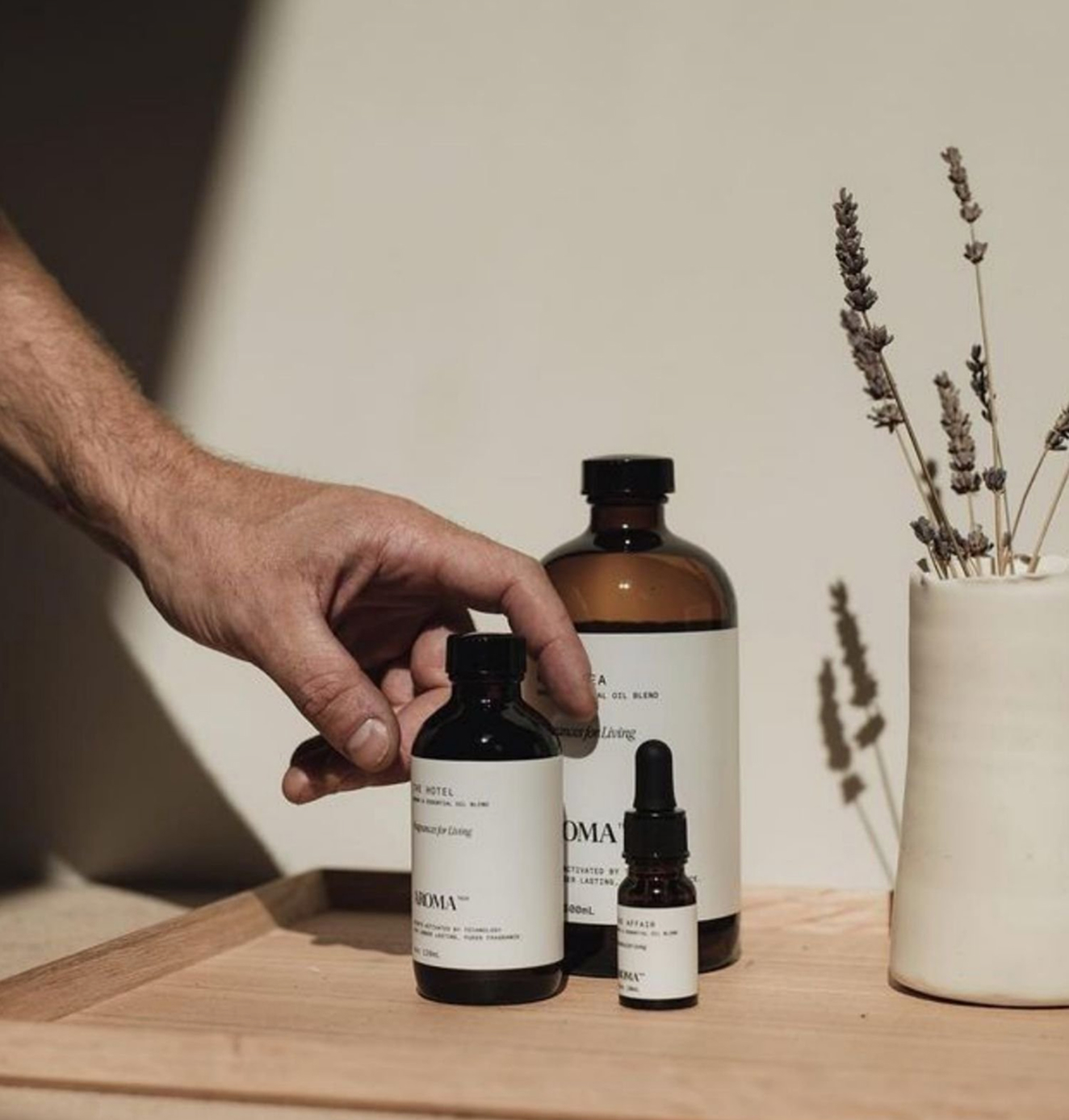 Hand arranging Aromatech essential oil bottles on a wooden tray beside a ceramic vase with lavender stems.