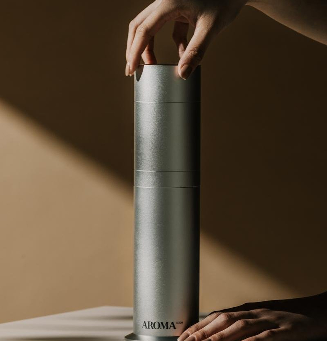 Hand placing the top lid on a tall, silver Aromatech diffuser against a warm-toned background.