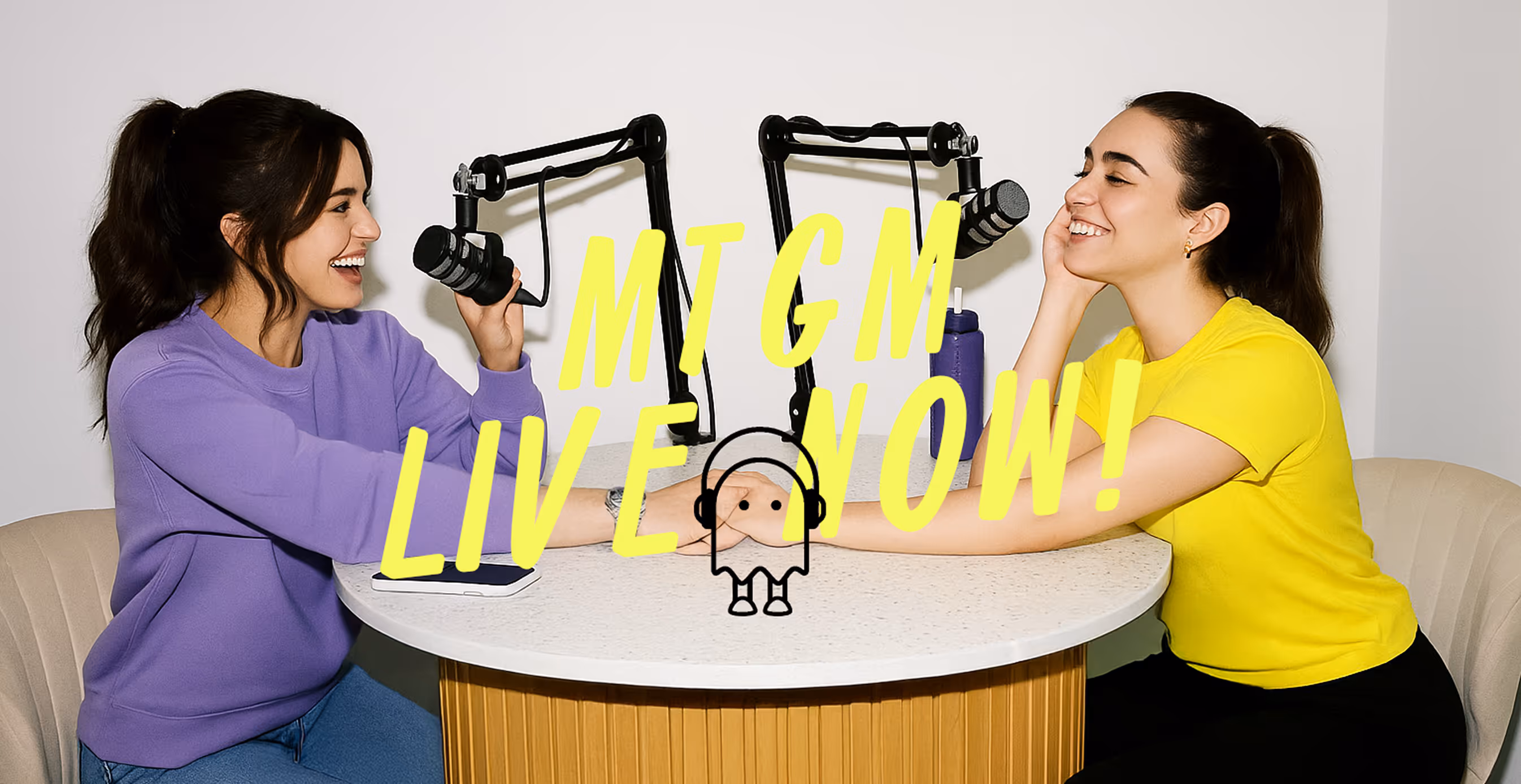 Two women recording a podcast at a table with microphones, smiling and holding hands, with bold yellow text overlay reading “MTGM LIVE NOW!”.