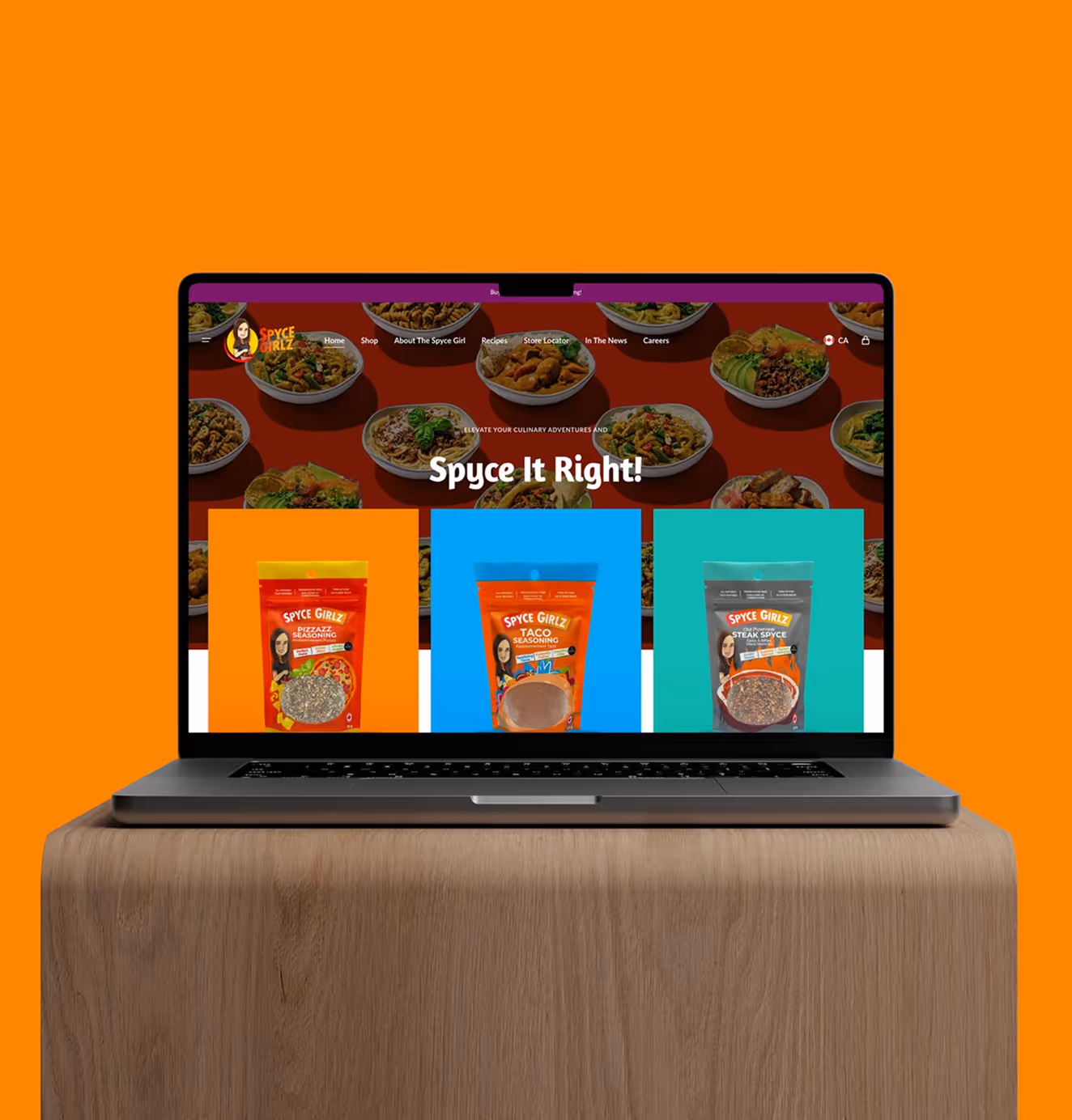 Laptop on a wooden surface displaying the Spyce Girlz website with bold colors, spice blend packaging, and the headline “Spyce It Right!” against an orange background.