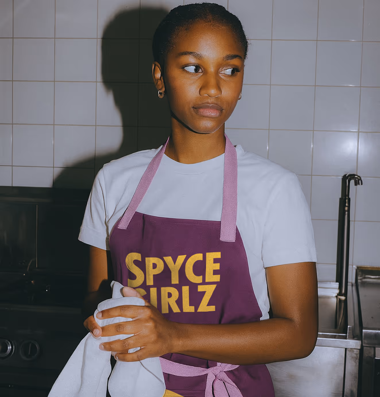 Young woman wearing a purple apron with yellow “Spyce Girlz” text, standing in a kitchen with white tiled walls, holding a dish towel.