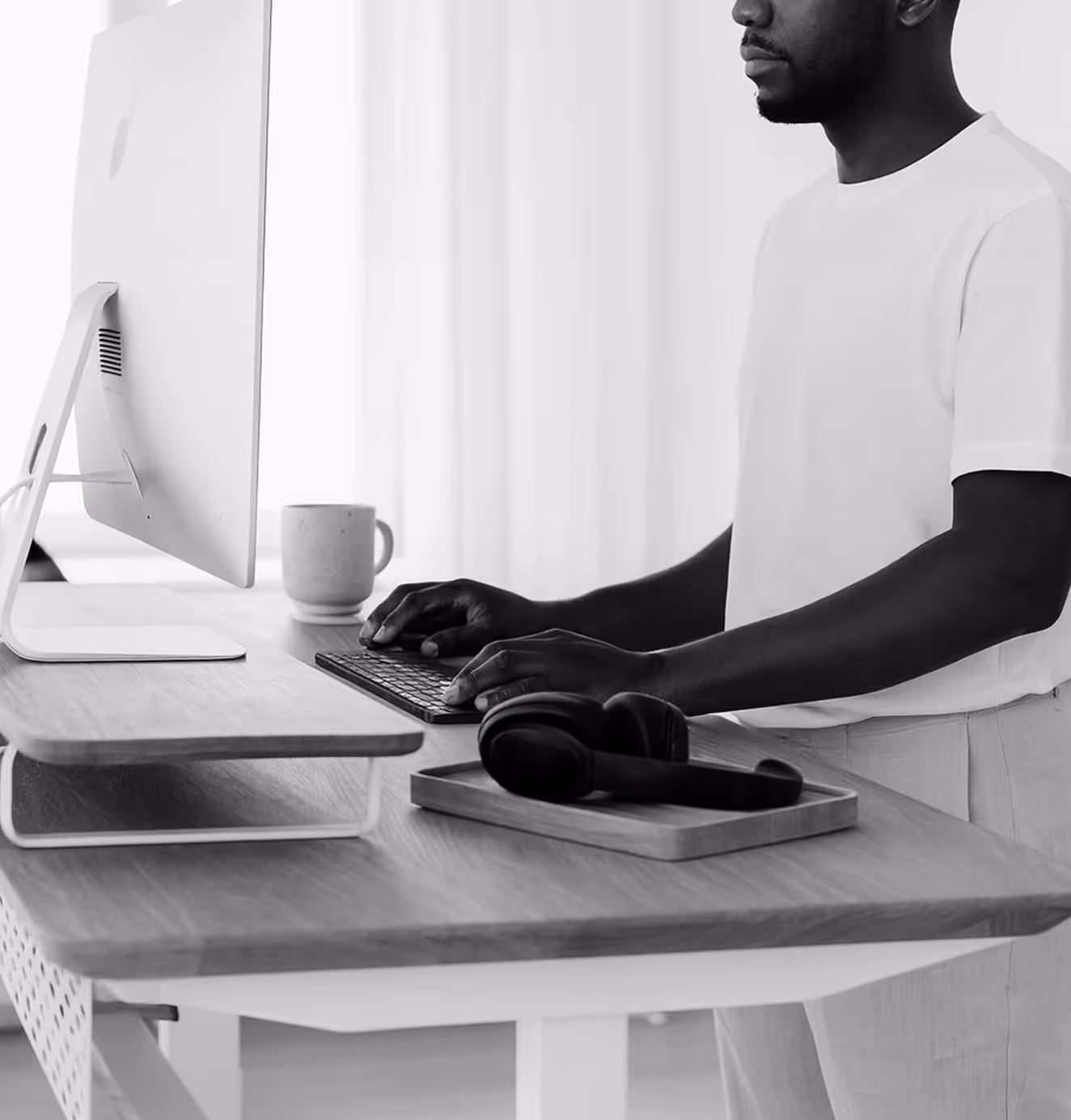 A man types at a modern standing desk with a monitor riser, mug, and headphones, representing HumanCentric’s ergonomic workspace design.