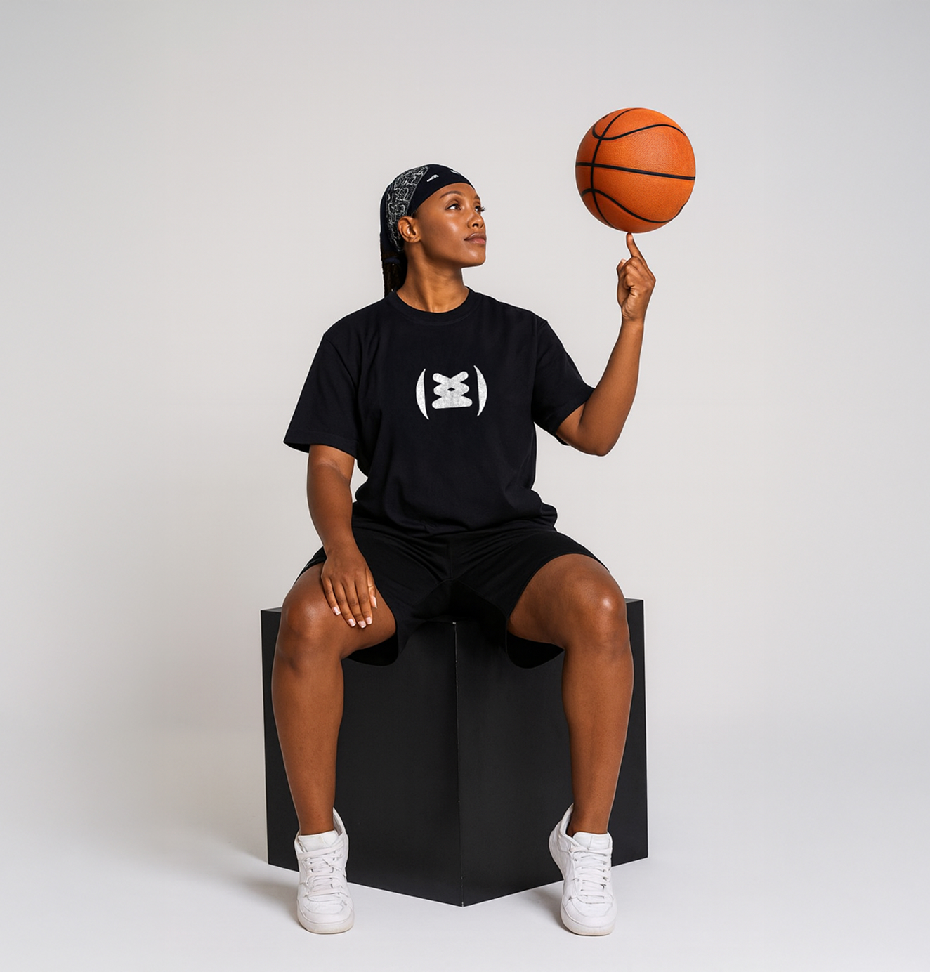 A woman in a black Sneakertopia t-shirt spins a basketball on her finger while seated on a cube in a studio.
