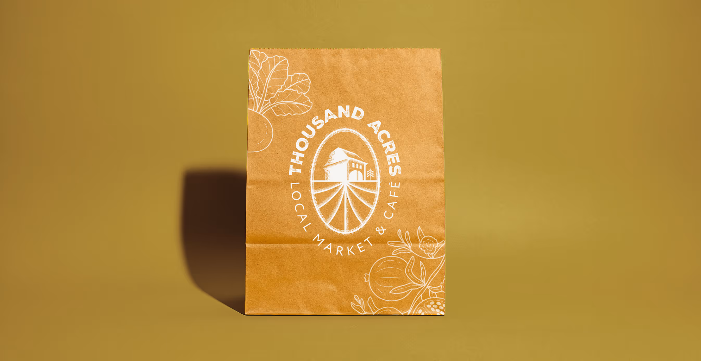 A brown paper grocery bag featuring the Thousand Acres Local Market & Café logo is placed against a mustard yellow background.