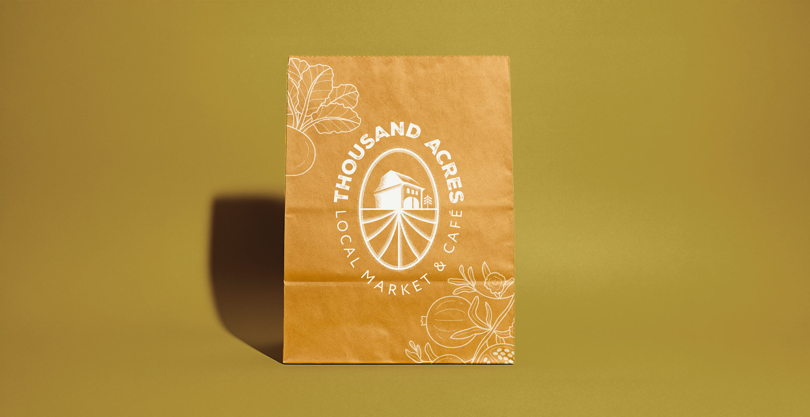 A brown paper grocery bag featuring the Thousand Acres Local Market & Café logo is placed against a mustard yellow background.