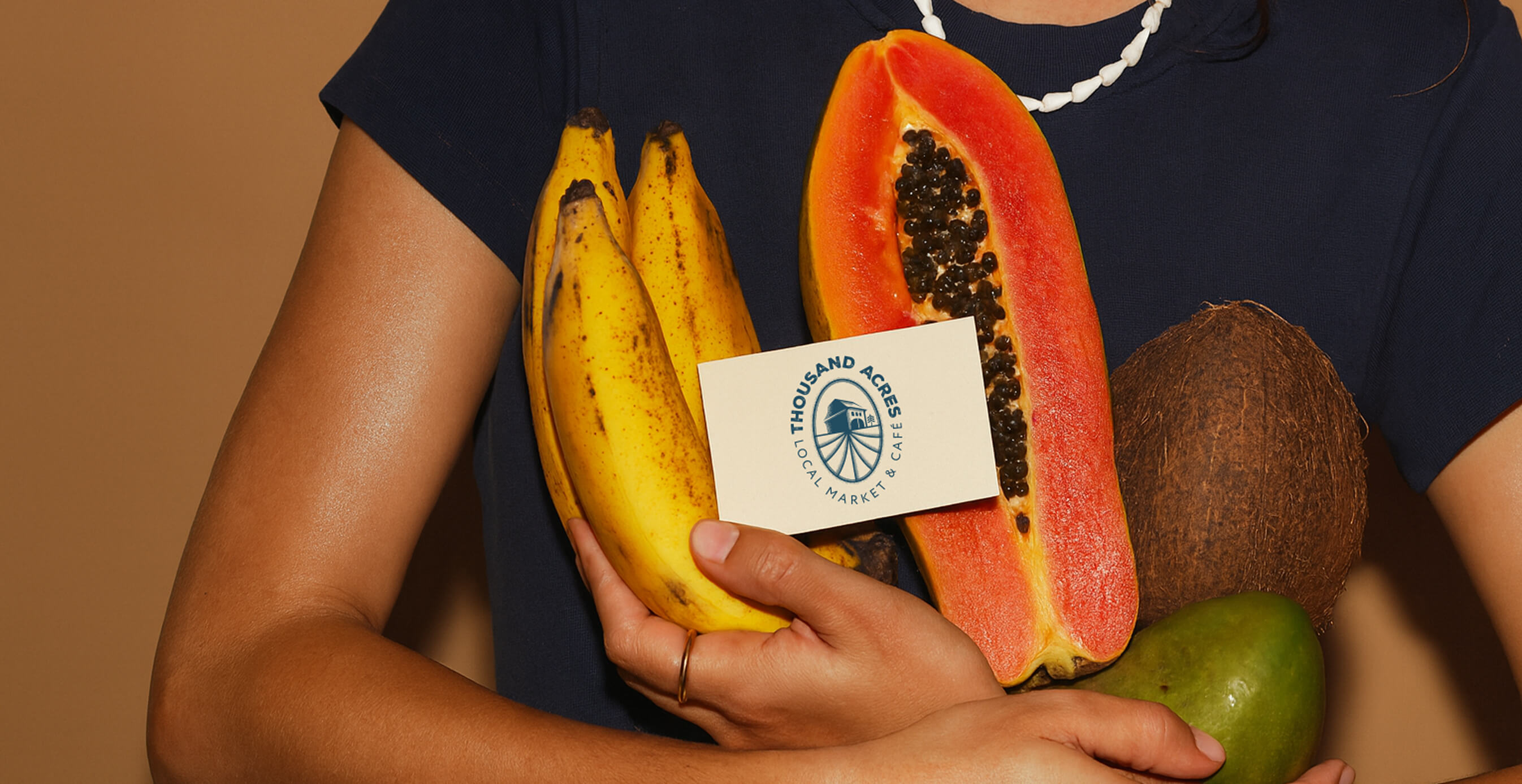 A person in a navy shirt holds tropical fruits including bananas, papaya, coconut, and mango, with the Thousand Acres logo card in hand.