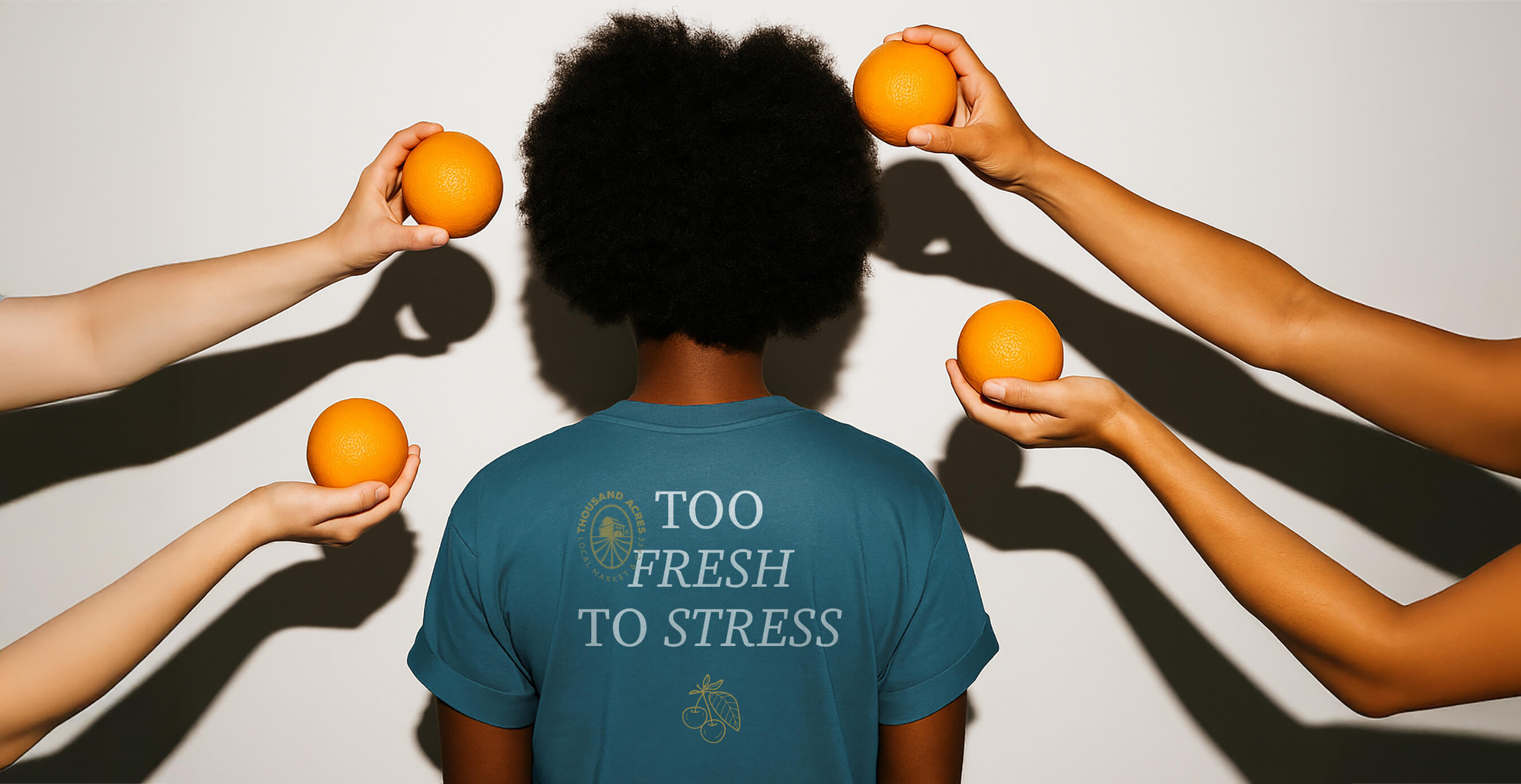 A person wearing a blue shirt printed with "Too Fresh to Stress" is surrounded by hands holding bright oranges against a white background.