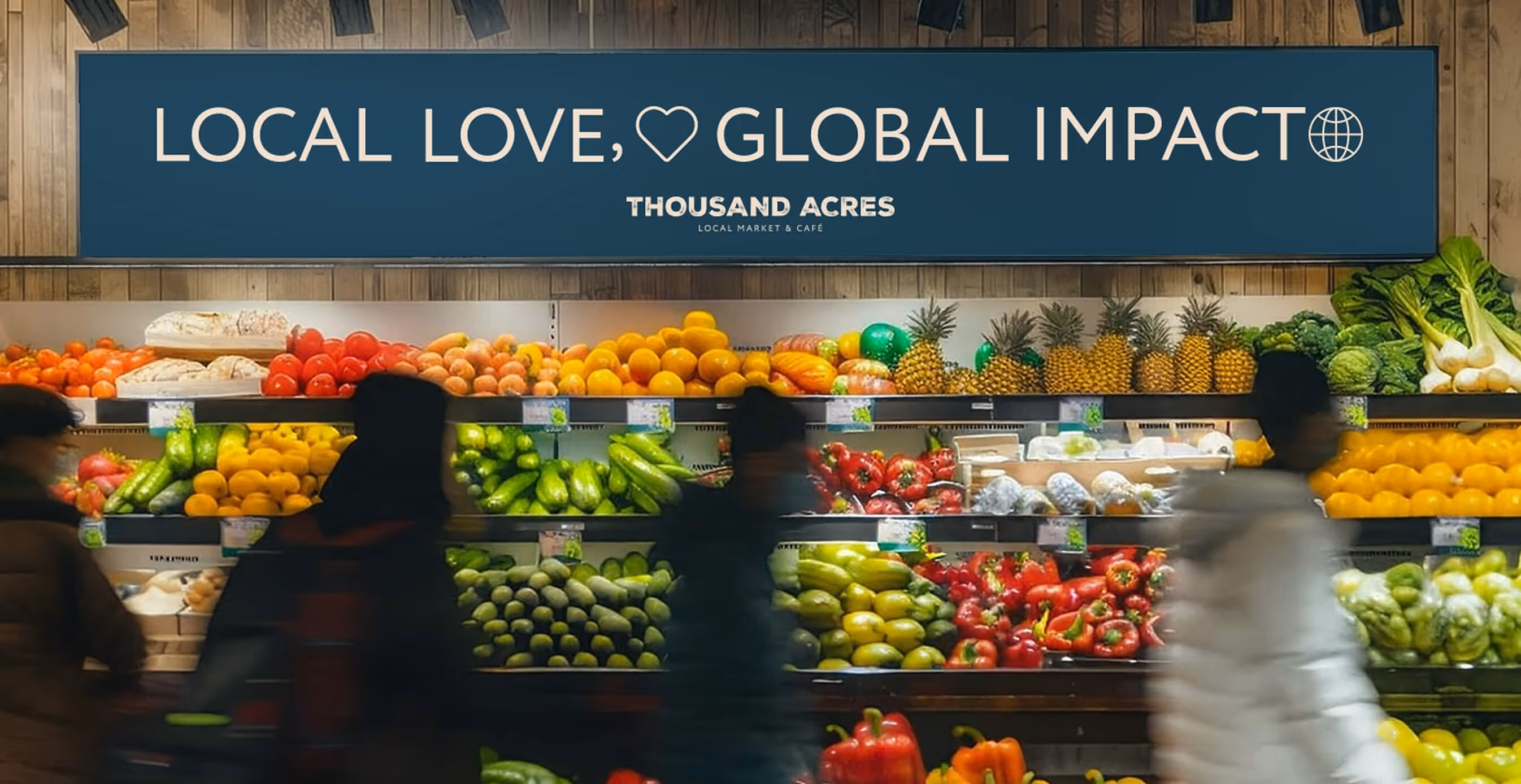 A grocery display at Thousand Acres with colorful fresh produce like tomatoes, cucumbers, peppers, and pineapples under a sign reading "Local Love, Global Impact."