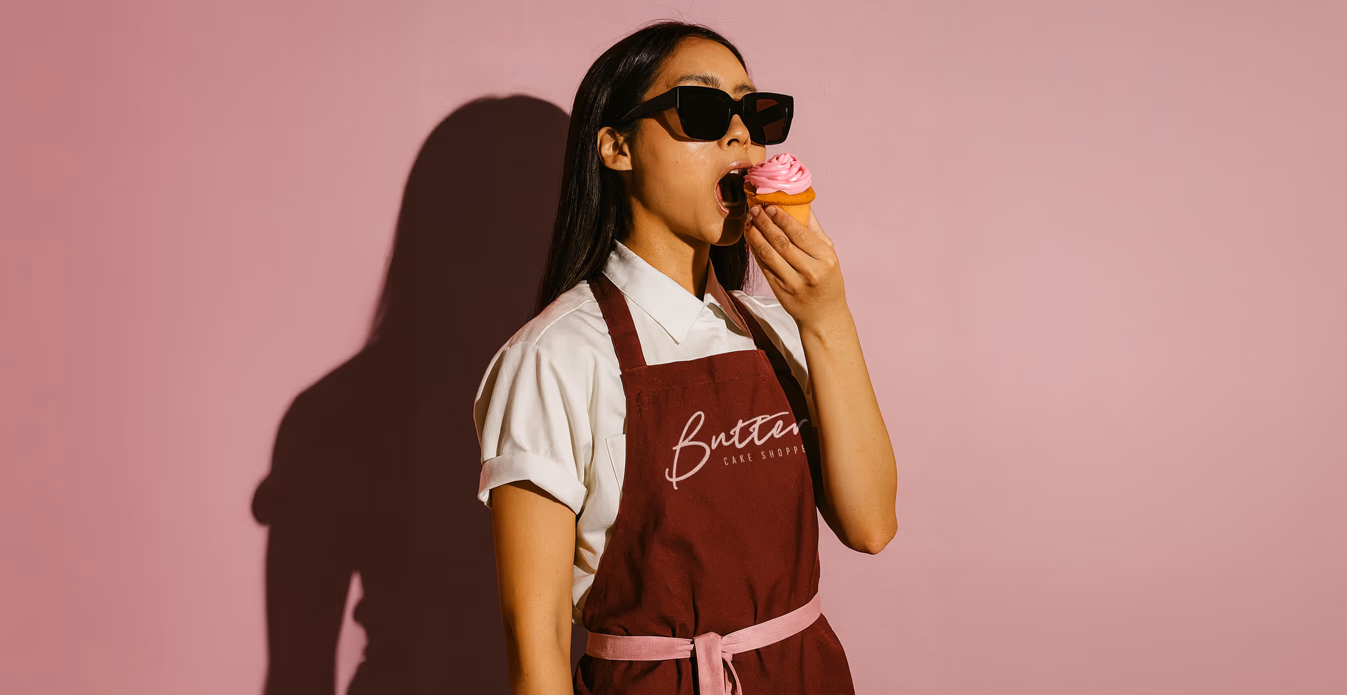 Woman wearing sunglasses and a burgundy apron with "Butter Cake Shoppe" logo takes a bite of a pink frosted cupcake against a pink background.